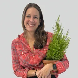 Blythe Yost, owner of Rockfield Landscape, wearing a red plaid button-up shirt with long brown hair smiles warmly at the camera while holding a potted evergreen plant. She is photographed against a white background and is wearing a dark-colored watch or fitness tracker on her wrist.